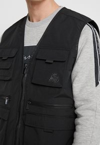 Svart zip-up väst med flera framsidans fickor, tillverkad av lättviktsmaterial. Har ett präglat logotyp och en slät grå sweatshirt under.