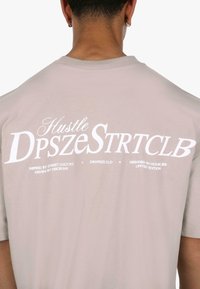 Lyse beige kortærmet t-shirt med stor hvid tekst på ryggen, der står "Hustle DpszeStrtClb" med yderligere mindre tekst nedenunder.