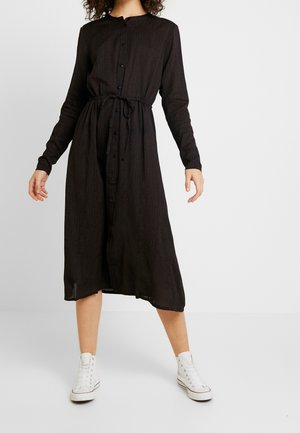 Czarna sukienka z długim rękawem, zapinana na guziki z przodu, z podkreśloną talią i luźnym krojem, zestawiona z białymi wysokimi trampkami.