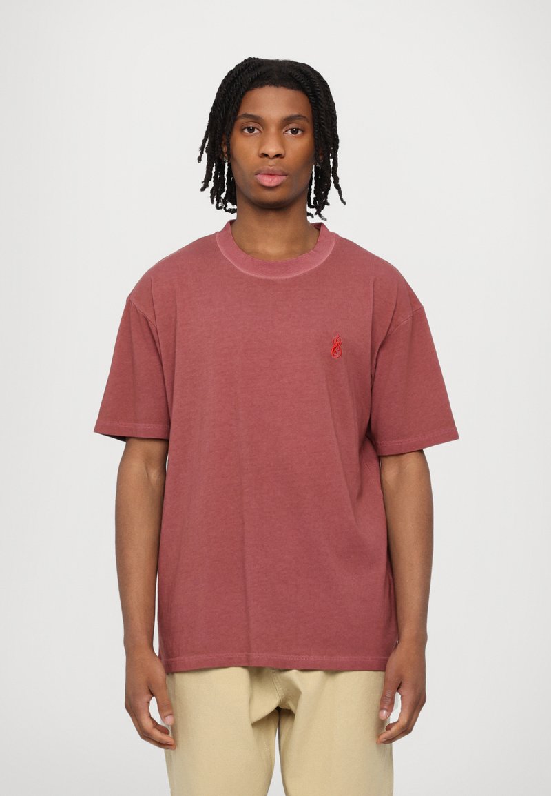 Camiseta de algodón marrón con mangas cortas, escote redondo y un pequeño logo de llama roja en el lado izquierdo del pecho. Textura suave y corte relajado.