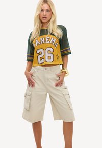 Femme blonde portant un maillot de sport court jaune et vert avec "PANEMA 26" et un short cargo beige mi-long.