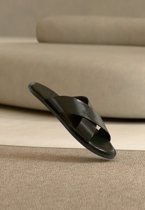 Schwarze Ledersandale mit gekreuzten Riemen auf beigem Teppich und Sofa im Hintergrund.