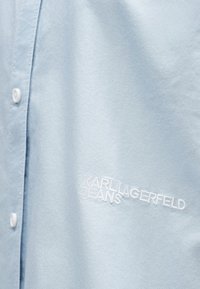 Ljust blå skjorta med vita knappar och broderad vit text "KARL LAGERFELD JEANS" på tyget.