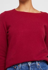 Bordeauxroter Pullover in Strickoptik mit weitem Ausschnitt und langen Ärmeln, kombiniert mit blauen, hoch taillierten Jeans.