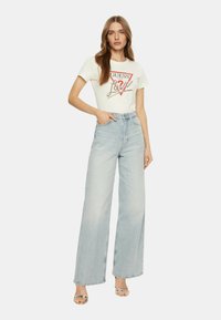 Weißes T-Shirt mit rotem und silbernem "GUESS"-Logo, kombiniert mit hellblauen, hochgeschnittenen, weiten Jeans und silbernen Absatzsandalen.