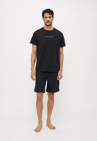 Crna pamučna majica s njuhom loga "Calvin Klein" u sredini, uparen s crnim kratkim hlačama koje imaju elastičnu pojase.