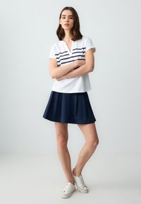 Weißes kurzärmliges Oberteil mit navyblauen horizontalen Streifen, kombiniert mit einem navyblauen ausgestellten Rock. Das Model trägt weiße Sneakers und steht mit verschränkten Armen.