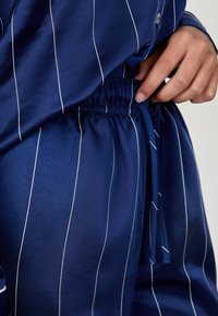 Granatowa satynowa bluzka od piżamy w białe pionowe paski, z elastycznym pasem na sznurku i gładką teksturą, podkreślająca elastyczny materiał.