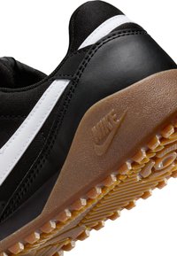 Zapatilla deportiva negra con parte superior de tela texturizada negra y acentos blancos, y una suela de goma color caramelo con patrones de dibujo prominentes.