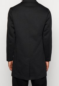 Cappotto lungo nero con un design su misura, caratterizzato da un taglio dritto, rever stretto e schiena con ventagli. Il tessuto appare liscio e opaco.