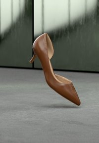 Καφέ δερμάτινο γυναικείο παπούτσι με ψηλό τακούνι και μυτερή μύτη, με λεπτό τακούνι, λείο υφή και ανοιχτή πίσω σχεδίαση, εμφανιζόμενο σε φόντο με απαλό χρώμα.