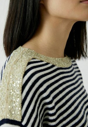 Épaule et cou d'une personne aux cheveux noirs et lisses portant un pull rayé noir et blanc avec une bordure en sequins dorés sur l'épaule et le col.