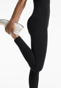 Leggings noirs en matériau extensible, design ajusté et texture lisse ; une main tient une sneaker blanche avec une semelle gris clair.