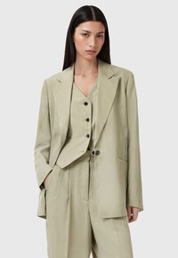 Completo di lino color khaki con giacca doppiopetto, rever a scanso, tre bottoni e pantaloni abbinati a vita alta con pieghe.