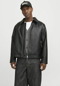 Black synthetic leather jacket with a pointed collar, front zipper closure, and side pockets, worn over a gray cardigan and dark jeans.