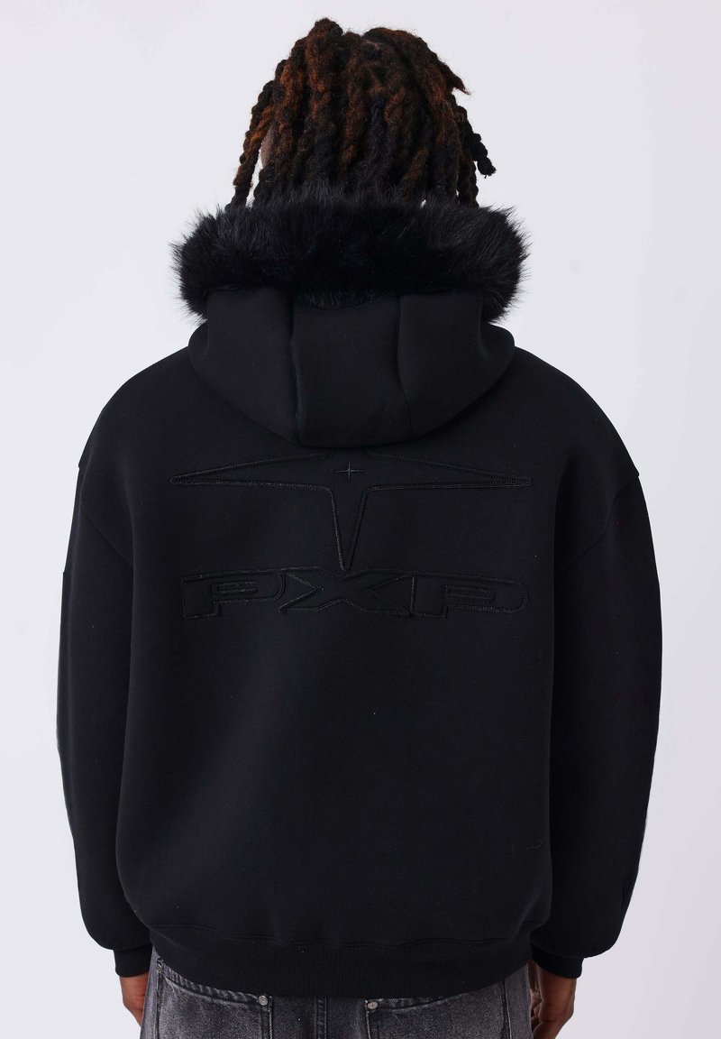 Personne aux cheveux foncés et bouclés portant un sweat à capuche noir avec une bordure en fourrure sur la capuche et un logo PXRD embossé dans le dos, vue de dos.