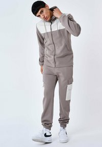 Hoodie in taupe met witte panelen, gecombineerd met bijpassende joggers met cargozakken en elastische boorden, gedragen met witte sneakers.