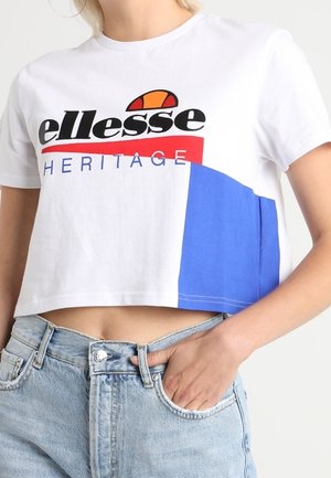 Vrouw draagt een witte cropped t-shirt met "ellesse Heritage" tekst en colour blocking ontwerp, gecombineerd met lichtblauwe hooggetailleerde jeans.