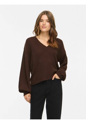 Femme portant un pull col V marron foncé à manches ballon et un pantalon noir, debout avec une main dans la poche sur un fond uni.