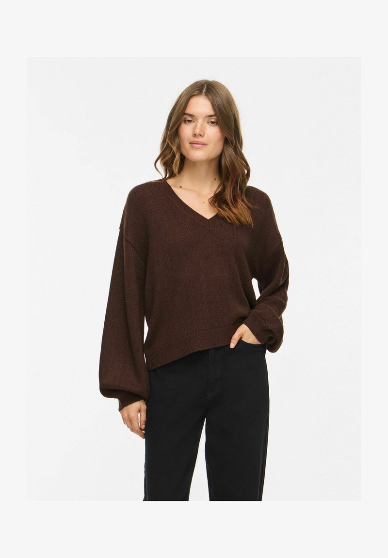 Femme portant un pull col V marron foncé à manches ballon et un pantalon noir, debout avec une main dans la poche sur un fond uni.