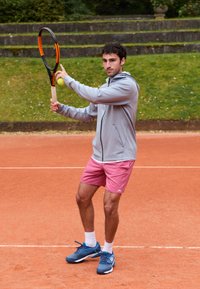 En man i grå luvtröja och rosa shorts förbereder sig för att serve med en tennisklubba och boll på en röd grus tennisbana.