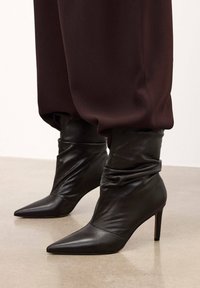 Melni ādas augstpapēžu potīšu zābaki ar smailiem pirkstiem un savilktu kāju daļu, ar gludu tekstūru un minimālistisku dizainu.