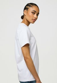 Hvid bomulds T-shirt med korte ærmer, rund halsudskæring og en afslappet pasform, vist på en model i profil. Glat tekstur, ingen mønstre.