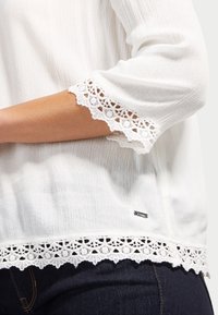 Biała bluzka z rękawami trzy czwarte, ozdobiona koronką przy mankietach i na dole. Materiał o fakturze oraz mały metalowy detal z logo na dole.