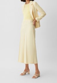 Jasnożółty garnitur z dopasowaną marynarką i szerokimi nogawkami spodni, zestawiony z beżowymi sandałami na paski. Gładka tkanina i subtelne detale z połyskiem.