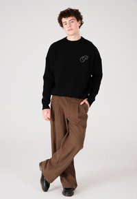 Junger Mann steht mit einer Hand in der Tasche, trägt einen schwarzen Pullover mit Logo, braune weit geschnittene Hose und schwarze Schuhe vor einem einfarbigen Hintergrund.
