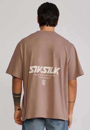 Mand iført brun oversized SikSilk t-shirt med hvidt logo og tekst på ryggen, kombineret med blå jeans, set bagfra.