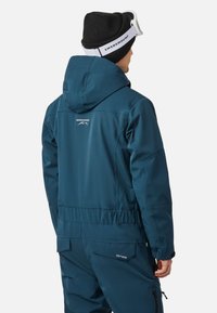 Teal waterdichte jas met een capuchon, elastische taille en logo-detail op de achterkant. Bevat een slank, geïsoleerd ontwerp dat geschikt is voor winterkleding.