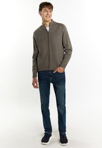 Maglione grigio con zip e una finitura testurizzata, indossato sopra una camicia bianca, abbinato a jeans blu e sneakers blu scuro. Il modello sta in piedi con un leggero sorriso.