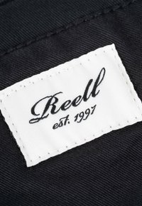 Schwarzes Stoffpatch mit weiß gesticktem Label, auf dem "Reell" in Kursivschrift und "est. 1997" darunter steht, wobei die Textur und die klare Typografie hervorgehoben werden.
