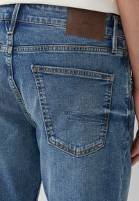 Blauwe spijkerbroek met een achterzak met stikdetails, een bruine merkpatch en een licht vervaagde textuur.