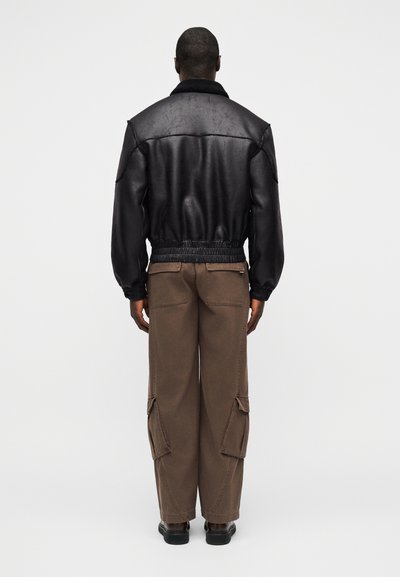 Άνδρας που φοράει μαύρο δερμάτινο μπουφάν bomber και καφέ παντελόνι cargo, στέκεται γυρισμένος με την πλάτη σε απλό λευκό φόντο.