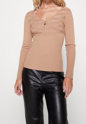 Femme portant un pull côtelé beige à manches longues avec un détail de fermeture éclair et un pantalon en cuir noir, debout devant un fond uni.