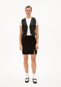 Frau in schwarzem, geknöpftem Weste über weißem T-Shirt, schwarzer Rock mit vorderem Schlitz, weißen Socken und schwarz-weißen Slippern vor weißem Hintergrund.