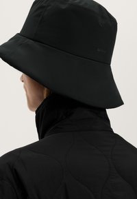 Person med en svart, bredbrättad bucket hat och en svart quiltad jacka, sedd bakifrån mot en neutral bakgrund.