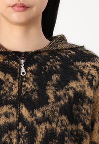 Sudadera de punto con estampado marrón y negro con un acabado texturizado, que cuenta con un cierre de cremallera completo y una capucha, destacando un llamativo diseño de estampado animal.