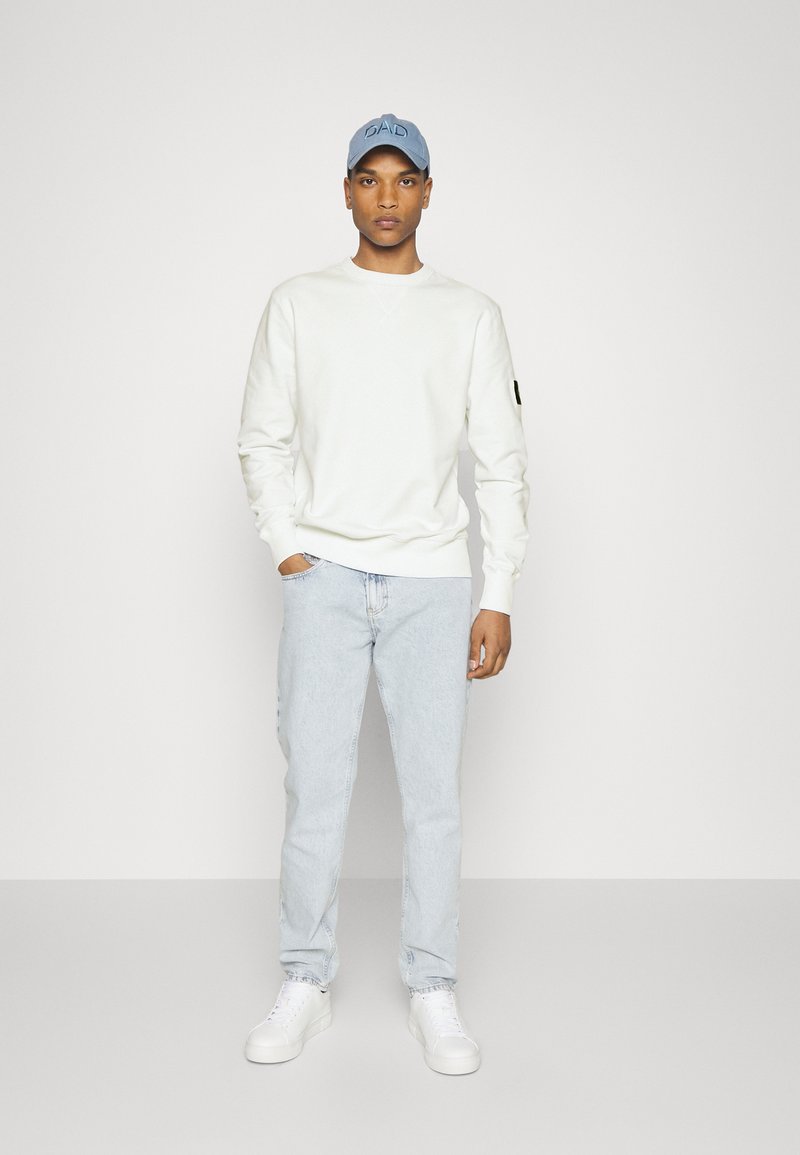 Felpa bianca con colletto rotondo, slim fit, e toppa sulle maniche; jeans azzurri; sneakers bianche; cappellino blu con testo "DAD".