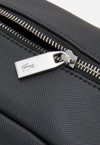 Svart teksturert stoffveske med en metallglidelås med krokodillogo og blankt sølvfarge hardware.