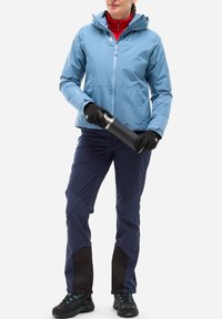 Femme portant une veste bleue, un pantalon marine, des gants noirs et des chaussures de randonnée tenant une bouteille d'eau noire avec les deux mains.