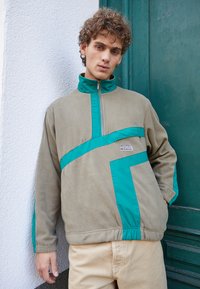 Sudadera de forro polar en beige con acentos en azul verdoso; cuello con media cremallera, patrón geométrico en la parte frontal, puños y cinturilla elásticos.