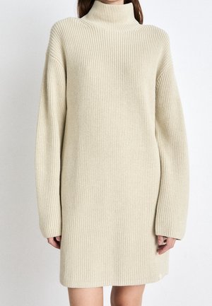 Femme portant une longue robe-pull en maille côtelée beige à col roulé avec des manches longues sur un fond uni.