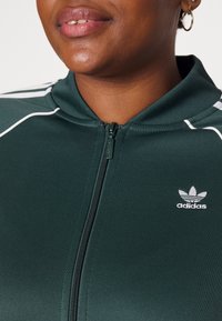 Grön zip-up jacka tillverkad av slät tyg, med vita ränder och en broderad vit Adidas-logotyp på vänster sida.