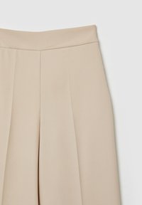 Jupe beige taille haute avec plis avant, tissu lisse et ceinture ajustée sur fond blanc.