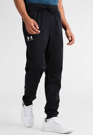 Mand iført sorte Under Armour joggingbukser og hvide sportssko står mod en ensfarvet baggrund.