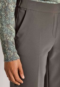 Linke Hand neben der Tasche einer grauen, maßgeschneiderten Hose, trägt einen Pullover mit Rippbündchen und einem abstrakten Muster in Blau, Grau und Beige.