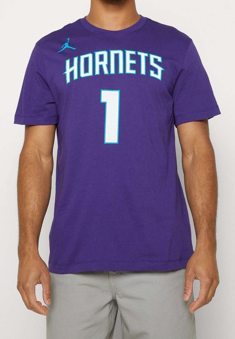 Homme portant un t-shirt violet à manches courtes avec ""HORNETS"" et le numéro 1 imprimés en bleu clair et blanc sur le devant.
